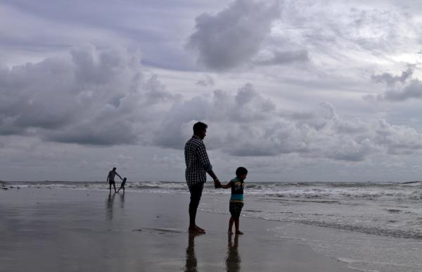 A boy holds his father's hand as they walk on a beach in the backdrop of pre-mo<em></em>nsoon clouds in Kochi