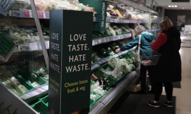 A shopper in a Waitrose fresh produce section with a sign that reads ‘Love taste, hate waste: choose loose fruit and veg’