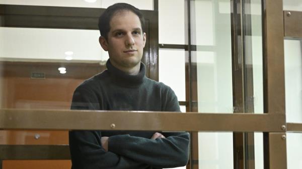 Evan Gershkovich stands behind a glass window in a room in court.