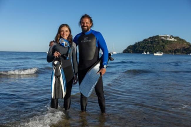 A man and a woman in diving gear stand in the surf on a beach