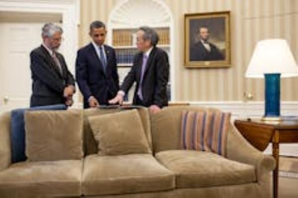 Three men stand over a couch discussing briefing material inside the White House Oval Office.