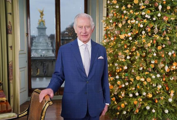 King Charles III delivering his Christmas address, dressed in a suit, standing in front of a Christmas tree at Buckingham Palace.