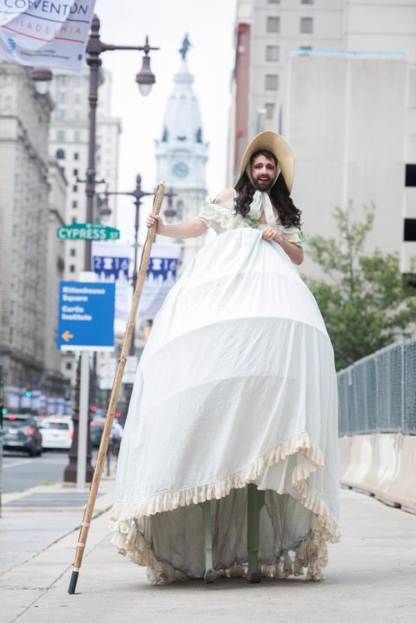 A member of the Bearded Ladies Cabaret, in a white dress and hat holding a stick, walking down Broad St. before a Bernie Sanders supporters march at the Democratic Natio<em></em>nal Convention, July 2016