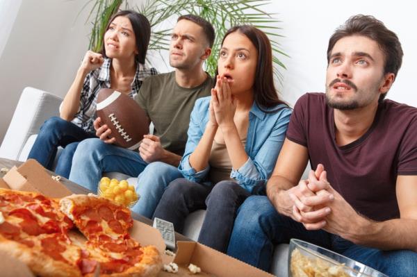 Group of young men and women intently watching a football game on TV, looking worried a<em></em>bout their team's performance