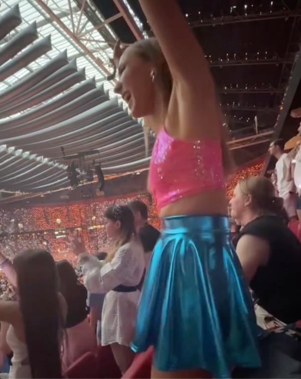 Nikki Glaser in a blue skirt and pink top dancing in a stadium at a Taylor Swift concert